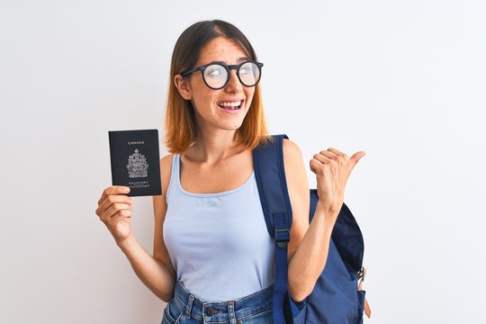 Beautiful redhead student woman wearing backpack and holding passport of canada pointing and showing with thumb up to the side with happy face smiling