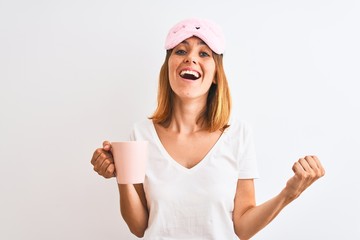 Beautiful redhead woman wearing sleeping mask drinking a cup of coffee over isolated background screaming proud and celebrating victory and success very excited, cheering emotion