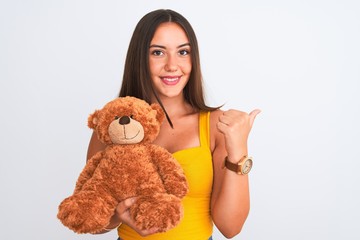 Young beautiful girl holding cute teddy bear standing over isolated white background pointing and showing with thumb up to the side with happy face smiling
