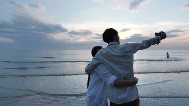 Happy Asian young couple in love use smartphone to take pictures of themselves and Spinning around together on the beach Background sunset in sea. Holiday rest and Travel concept. Slow Motion