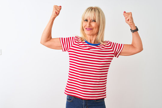 Middle age woman wearing casual striped t-shirt standing over isolated white background showing arms muscles smiling proud. Fitness concept.