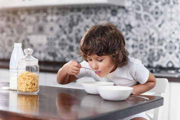 Happy little boy eats with pleasure healthy breakfast of cornflakes and milk at the kitchen.