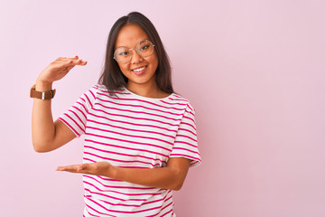 Young chinese woman wearing striped t-shirt and glasses over isolated pink background gesturing with hands showing big and large size sign, measure symbol. Smiling looking at the camera. Measuring 