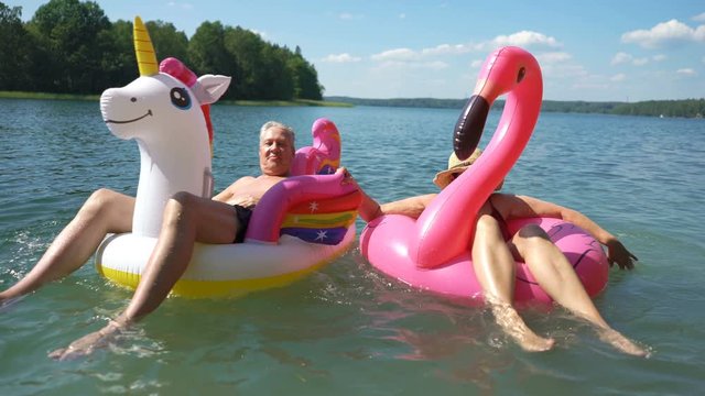 Elderly couple floating on inflatable flamingo and unicorn. Funny active pensioners happy together enjoying summer vacation on the beach in Europe, laughing, playing the fool, splashing water