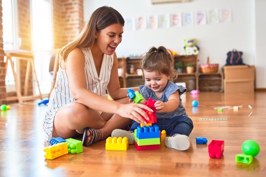 Young beautiful teacher and toddler playing with building blocks toy at kindergarten