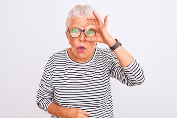 Senior grey-haired woman wearing striped navy t-shirt glasses over isolated white background doing ok gesture shocked with surprised face, eye looking through fingers. Unbelieving expression.
