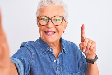 Grey-haired woman wearing denim shirt and glasses make selfie over isolated white background surprised with an idea or question pointing finger with happy face, number one
