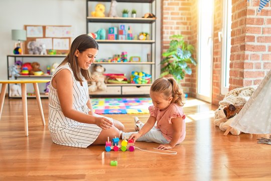 Beautiful teacher and blond student toddler girl playing with wooden train at kindergarten