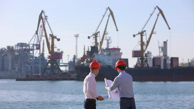 Handshake of architects in orange helmets on the background of the shipyard with port cranes
