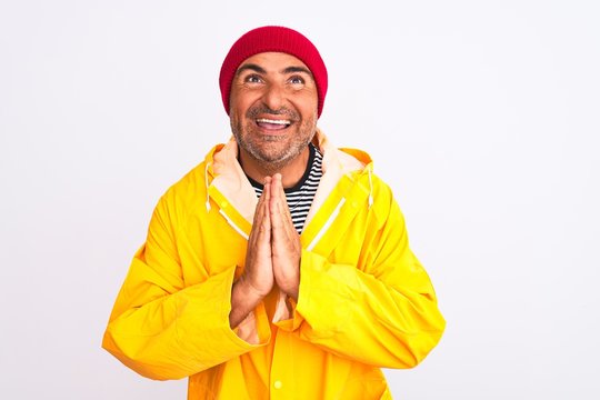 Middle age man wearing rain coat and woolen hat standing over isolated white background praying with hands together asking for forgiveness smiling confident.