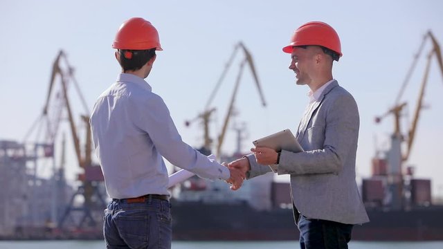 Two design engineers in helmets communicate and show the project on the tablet on the background of a dock with cargo port cranes