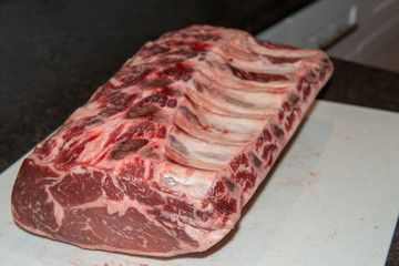 In the restaurant, a large prime rib roast sits on a white plastic cutting board.  The raw meat or beef has six ribs in the roast. Some spots of blood are on the cutting board.