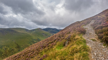 A scenic view of a mountain trail track along a grassy mountain slope and valley with mountain range in the background under a stormy grey cloudy sky