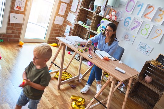Young caucasian child playing at playschool with teacher. Young woman sitting on the desk of the classroom