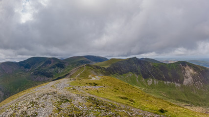 A scenic view of a rocky mountain summit with grassy green slope, trail and mountain range in the background under a stormy grey cloudy sky