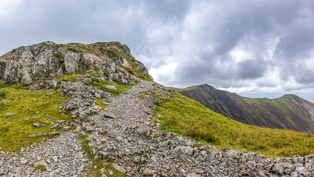 A scenic view of a rocky mountain summit with grassy green slope, trail and mountain range in the background under a stormy grey cloudy sky