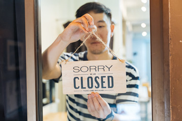 Through glass view of Asian male business owner hanging up sign with inscription closed at end of...