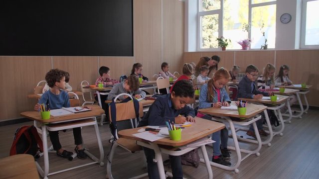 Primary school students sitting at desks and drawing while teacher walking between rows and helping little artists. Friendly asian woman interested in success of pupils in drawing during art lesson