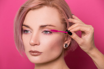 Beautiful blonde girl of European appearance holds in her hand a eyelash brush near her eyes isolated on a pink background