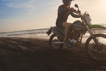 Male masculine model with cross road motorbike at the ocean coast with black sand at the golden hour time