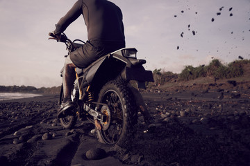 Male masculine model with cross road motorbike at the ocean coast with black sand at the golden hour time