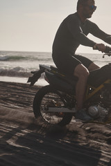 Fototapeta na wymiar Male masculine model with cross road motorbike at the ocean coast with black sand at the golden hour time
