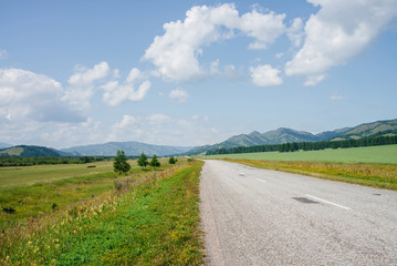 Fototapeta na wymiar Wonderful scenic landscape with long asphalt road through green fields with mountain view on horizon. Vivid green scenery with highway and meadow under blue sky in sunny day. Shiny asphalt in sunlight