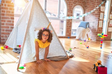 Beautiful toddler wearing glasses and unicorn diadem sitting on the floor inside tipi at kindergarten