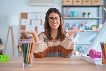 Young beautiful teacher woman wearing sweater and glasses sitting on desk at kindergarten smiling...