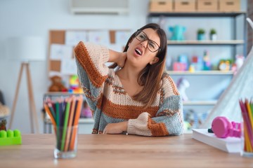 Young beautiful teacher woman wearing sweater and glasses sitting on desk at kindergarten Suffering of neck ache injury, touching neck with hand, muscular pain