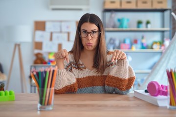Young beautiful teacher woman wearing sweater and glasses sitting on desk at kindergarten Pointing...