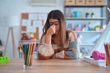 Young beautiful teacher woman wearing sweater and glasses sitting on desk at kindergarten tired...