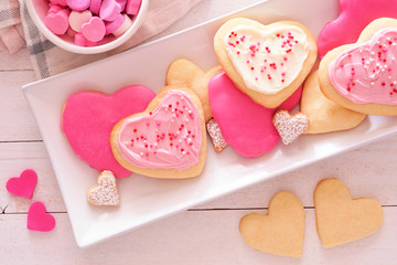Heart shaped Valentines Day cookies with pink and white icing and sprinkles. Overhead view on a serving plate against a white wood background.