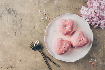 Pink mousse cakes in the shape of a heart decorated with mini hearts on a beige rustic background. Heart shaped cakes for Valentine's Day or Mother's day. Top view. Flat lay