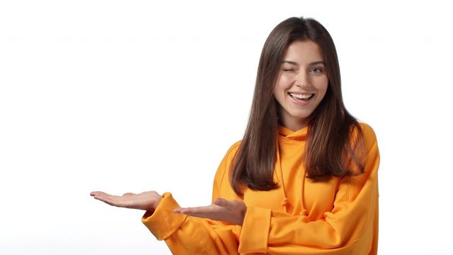 Slow-motion adorable cheeky caucasian woman in stylish hoodie, presenting product, introduce company banner over blank copy space, smiling wink, show thumbs-up in approval, recommendation