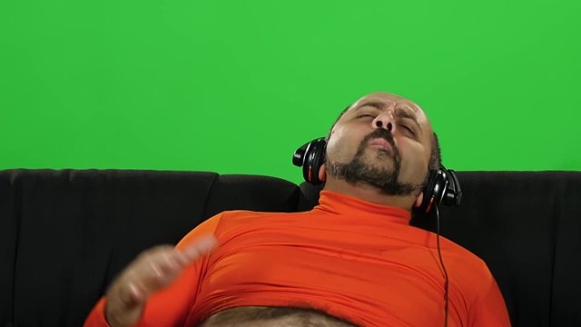 Funny fat caucasian guy dancing when sitting on sofa in big headphones. Green screen. Slow motion