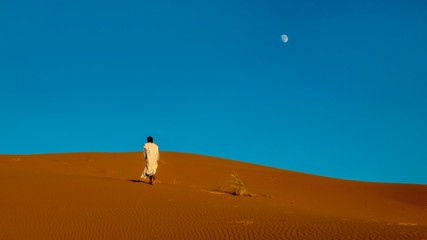 Fototapeta na wymiar A Moroccan man wearing full length traditional nomadic clothing, walks barefoot uphill toward the ridge of a red sand dune in the Sahara Desert.