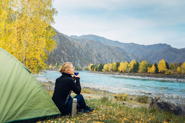 Young woman sits on grass near tourist tent and drinks hot tea/coffee from thermos cup on the background of river and mountains. Traveler resting after a hike, enjoying solitude and beauty of nature.