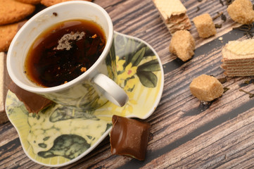 A Cup of black tea, tea leaves, pieces of brown sugar, oatmeal cookies, waffles, pieces of chocolate on a wooden background. Close up.