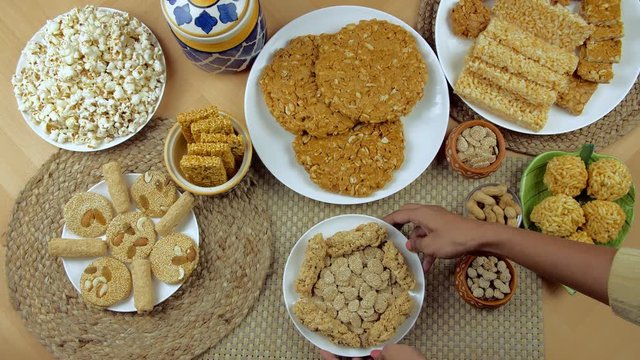 Hands of an Indian woman placing a plate of rewri and gajak on a wooden table - Lohri festival. Top view shot of beautifully plated Lohri items like til patti  peanuts chikki  rewri  gajak rolls  p...