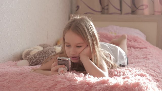 Cute girl playing smartphone lying on her bed in her room.