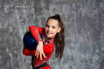 A portrait of young girl gymnast in a red suit makes exercise with a ball against a gray wall. She...