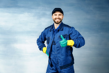 Thumb up. Young handsome man with a beard in a blue working uniform for cleaning rooms smiles...