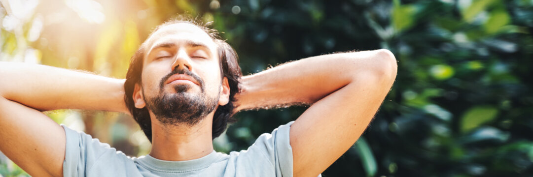 A bearded man is meditating outdoor in the park with face raised up to sky and eyes closed on sunny summer day. Concept of meditation, dreaming, wellbeing healthy lifestyle