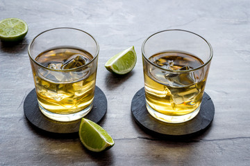 Whiskey with ice. To glasses of strong alcohol drink near lime on grey stone background