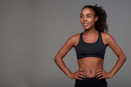 Positive young pretty sporty dark skinned woman with long brown curly hair looking cheerfully aside and keeping hands on her waist, isolated over grey background in athletic wear