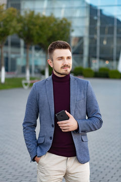 Portrait of handsome smiling man in casual wear holding smartphone. Young businessman analyzes the results of important phone conversation. Young man standing on the city street near business center.