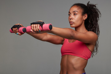 Concentrated young slim dark skinned curly woman with ponytail hairstyle working out her arms and looking in front of herself with folded lips, standing against grey background