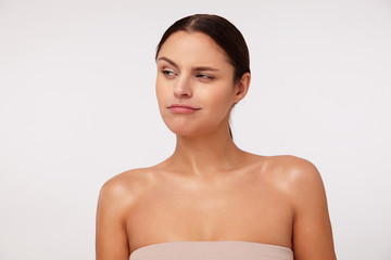 Indoor shot of lovely young brunette woman with casual hairstyle squinting and raising eyebrow while looking aside cunningly, standing over white background in beige top