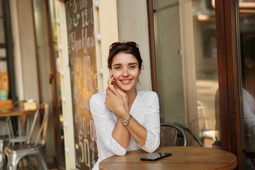 Shot of young beautiful brown-eyed brunette woman with sunglasses on her head keeping hands on table while looking cheerfully at camera, dressed in white polka-dot wear